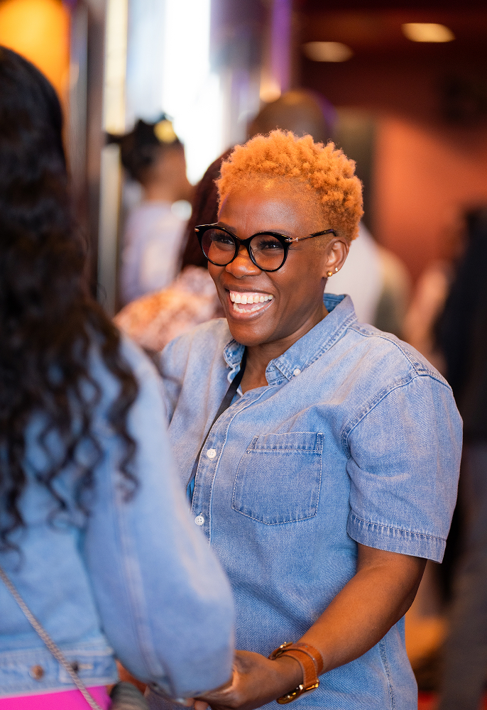 Smiling woman with short blonde hair and glasses wearing a denim shirt, holding hands with another person.