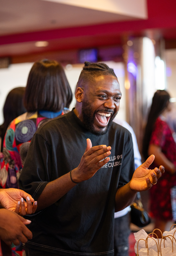 Man with braided hair laughing and clapping his hands at an indoor social event.