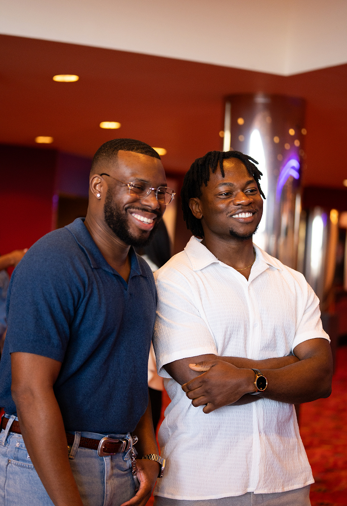 Two smiling men standing indoors with warm lighting, one in a navy polo shirt and glasses, the other in a white short-sleeve shirt with arms crossed.