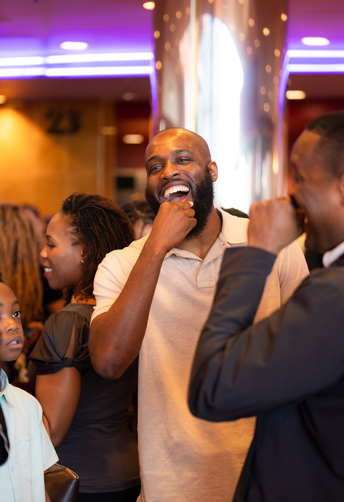 A group of people socializing indoors, with a man in a beige polo shirt laughing and playfully covering his mouth.