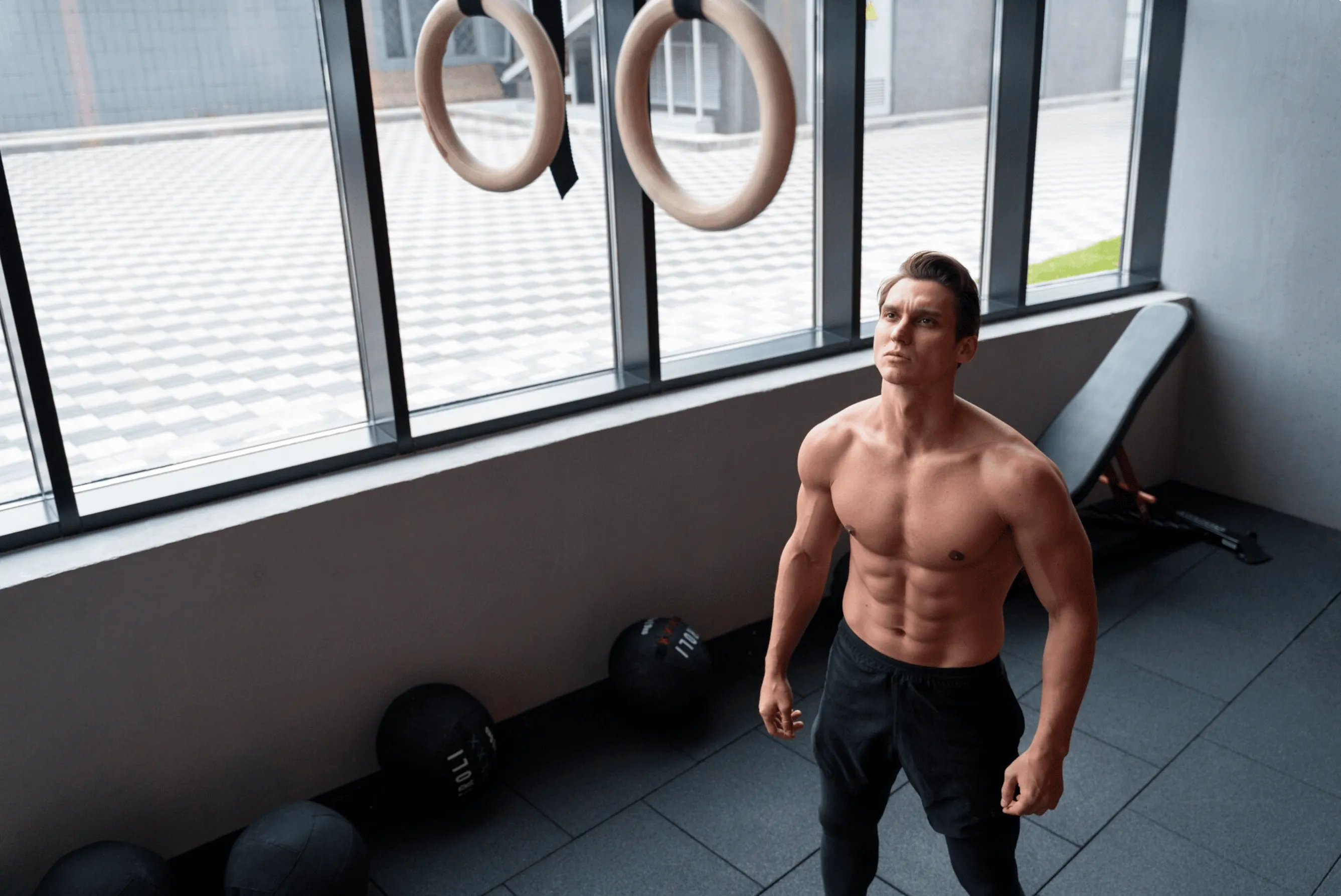 Muscular man standing in a gym beneath gymnastic rings, representing muscle maintenance and strength training supported by the best peptides for muscle maintenance.