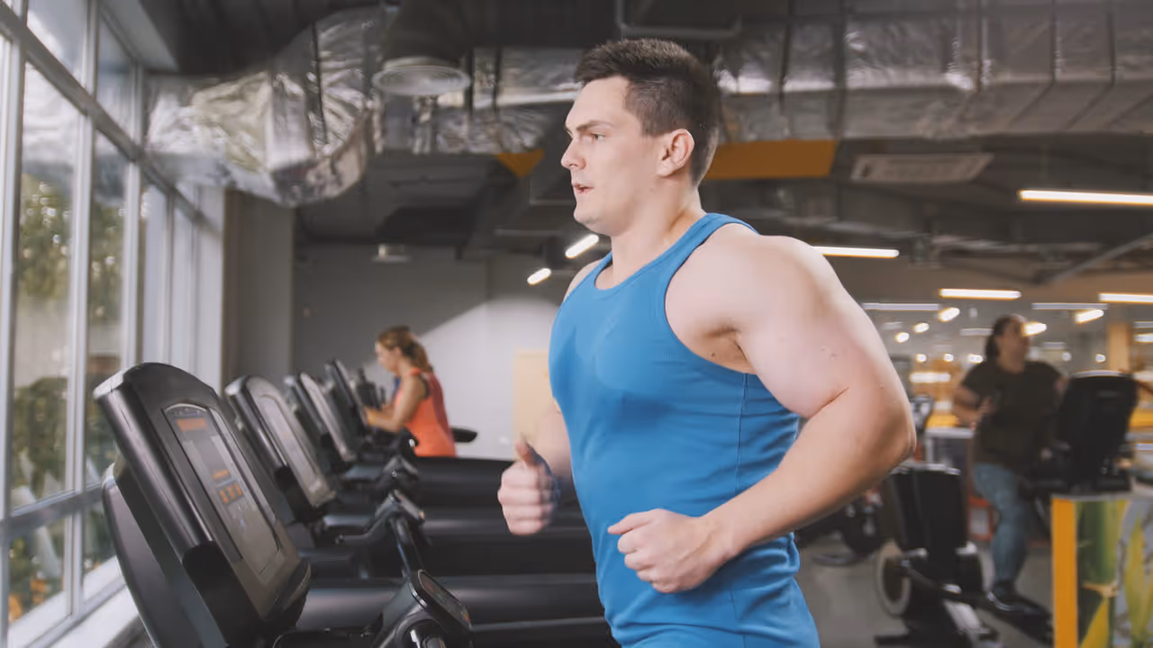 A man in a blue tank top runs on a treadmill, focused on weight loss amidst others exercising.