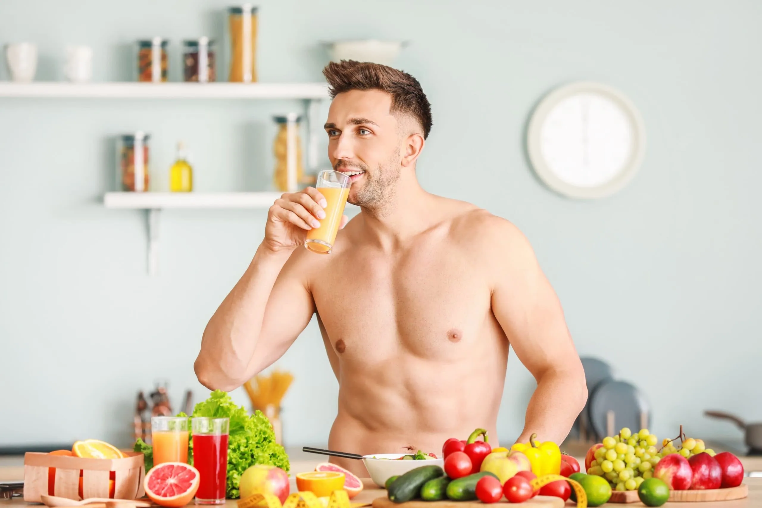 Shirtless man drinking a glass of juice surrounded by fresh fruits and vegetables, wondering if intermittent fasting has any effect on testosterone.