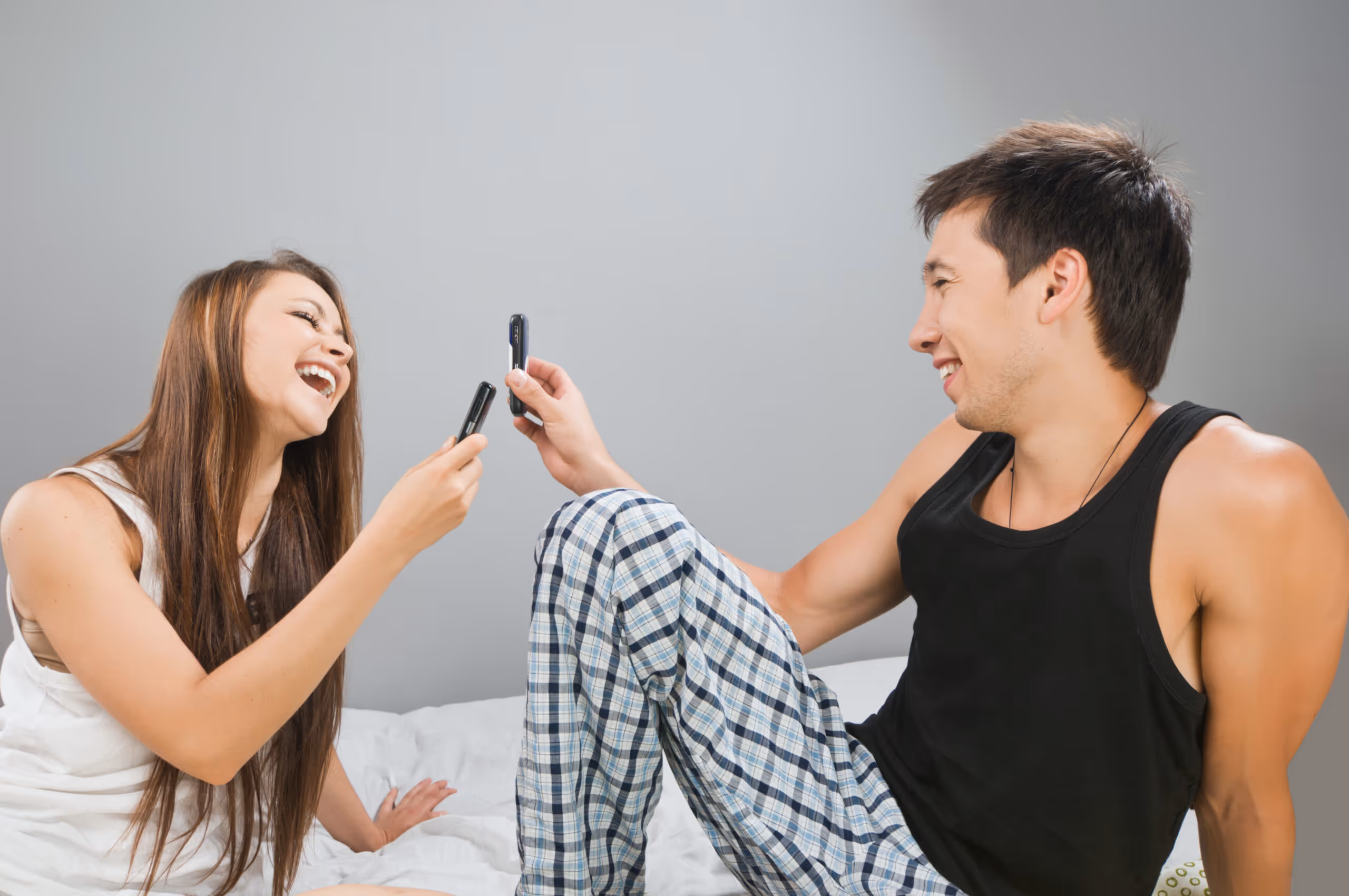 Smiling couple sitting on a bed and playfully comparing small devices, symbolizing intimacy, attraction, and the use of male sex drive enhancers to support libido.