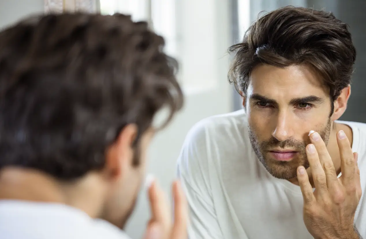 Man examining acne-prone skin in mirror, representing testosterone replacement therapy (TRT) and its impact on breakouts.