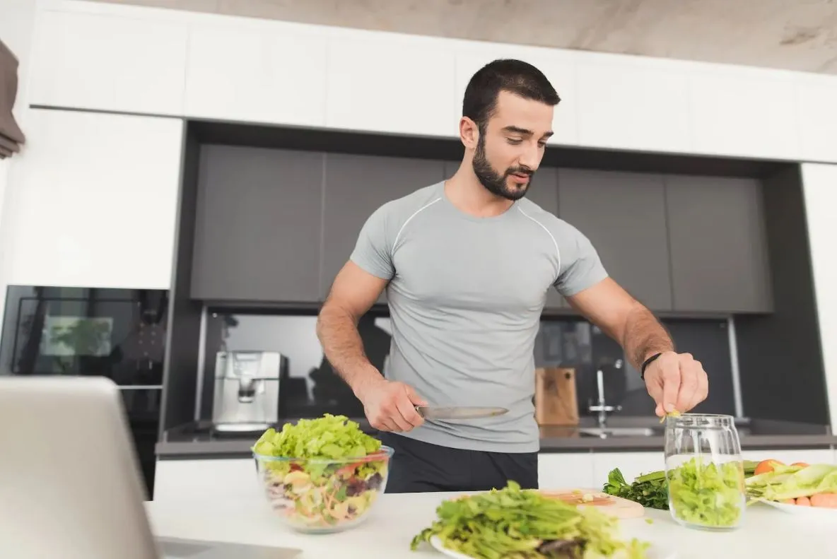 Fit man preparing a fresh salad in a modern kitchen, emphasizing healthy eating and lifestyle for energy and hormone balance.