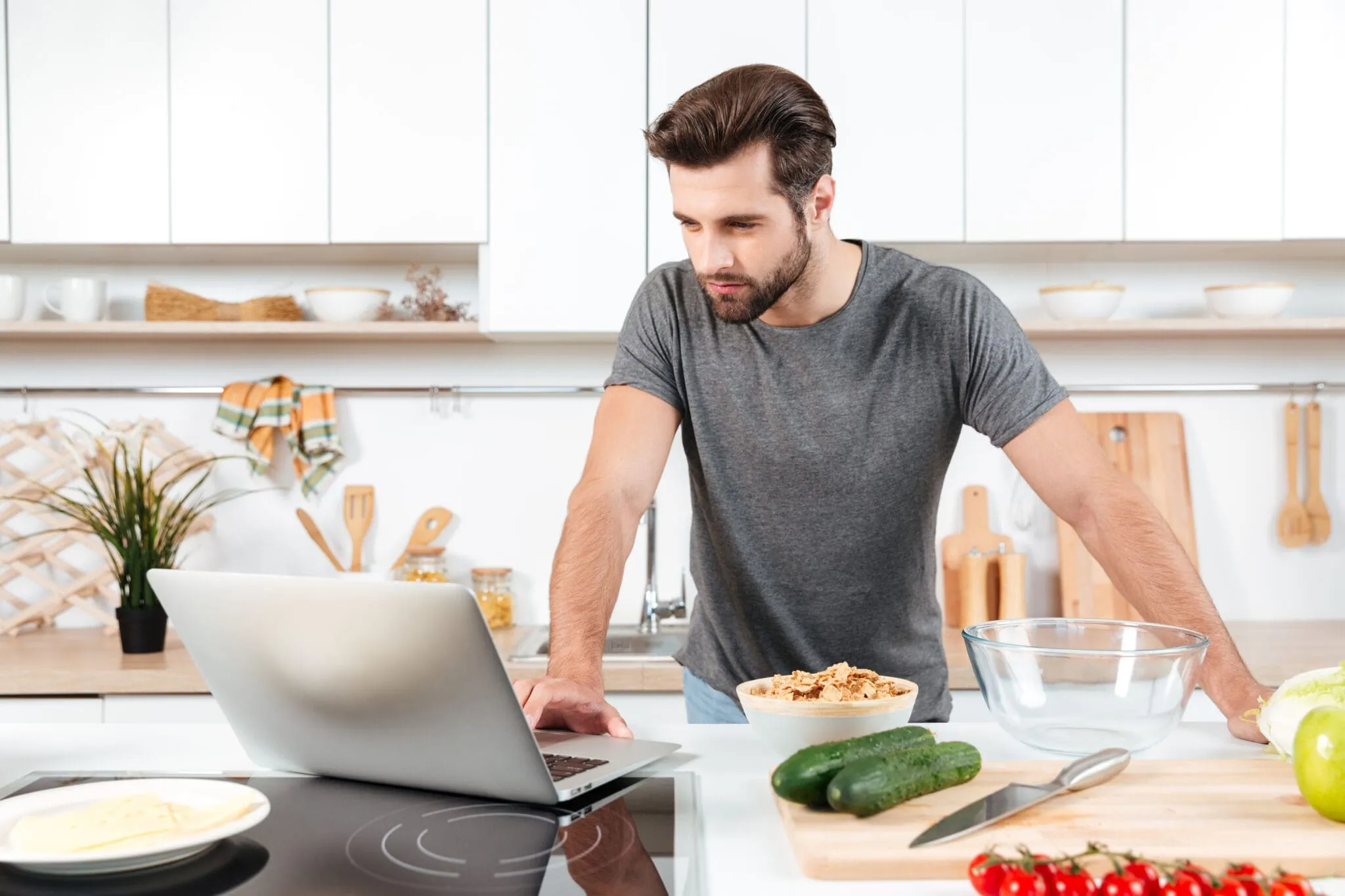Man preparing a healthy meal in a modern kitchen while looking at a recipe on a laptop, with fresh vegetables and cereal on the counter.