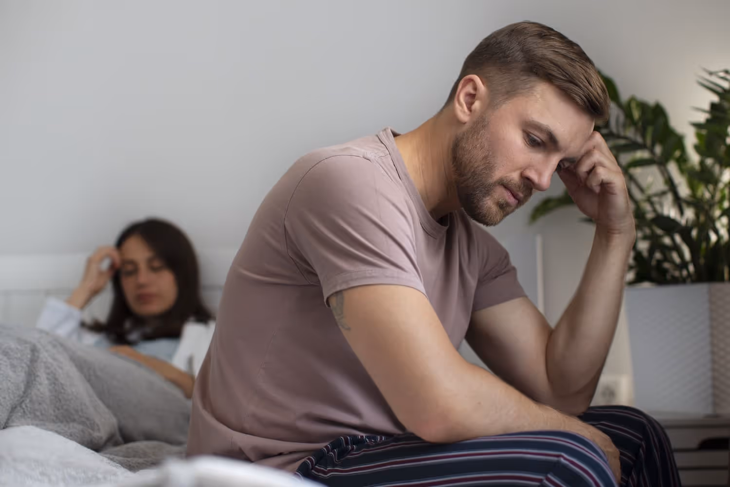 A young man looks stressed while sitting on the edge of a bed, reflecting on the challenges of performance anxiety, a common issue related to erectile dysfunction in younger men.