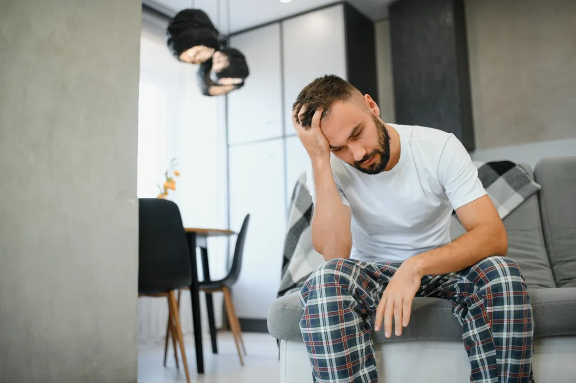 Man sitting on a couch in pajamas with his head in his hand, appearing fatigued or distressed, representing symptoms of male hypogonadism.
