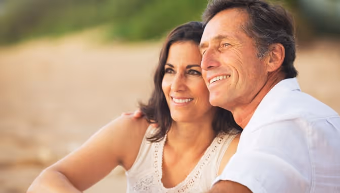 An older couple smiling and enjoying a peaceful moment together on a beach, symbolizing the positive results of treatments like GAINSWave® for enhancing men's health and vitality.