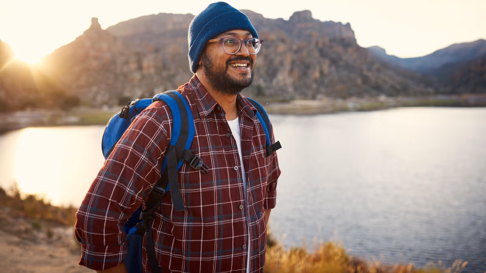 Smiling man with glasses and a backpack standing outdoors by a lake, symbolizing the idea of a healthy, active lifestyle that can be supported by hormone pellet therapy for men.