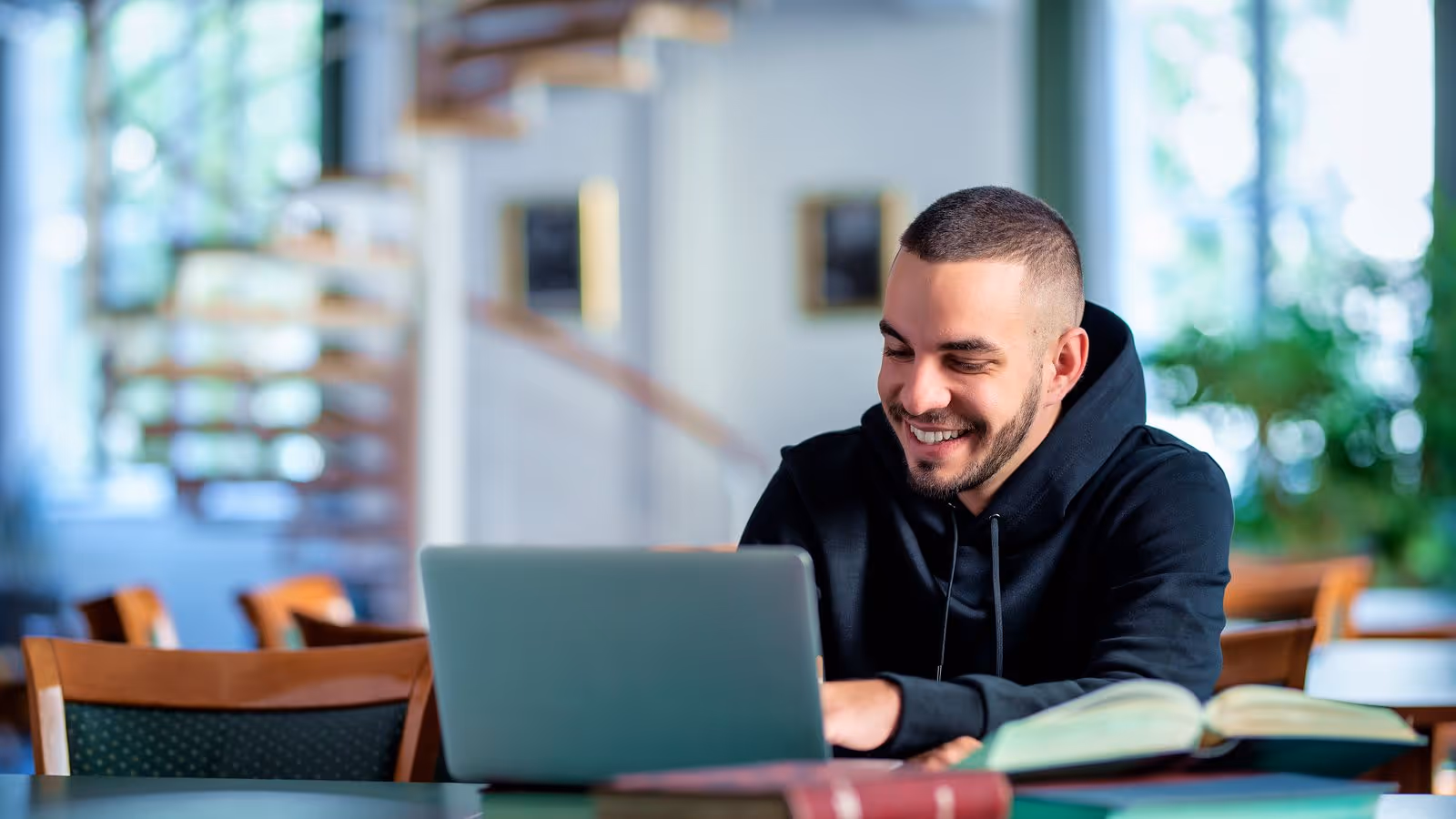 Smiling college student working on his laptop in a library, symbolizing the importance of essential vitamins for boosting health, energy, and performance in college men.