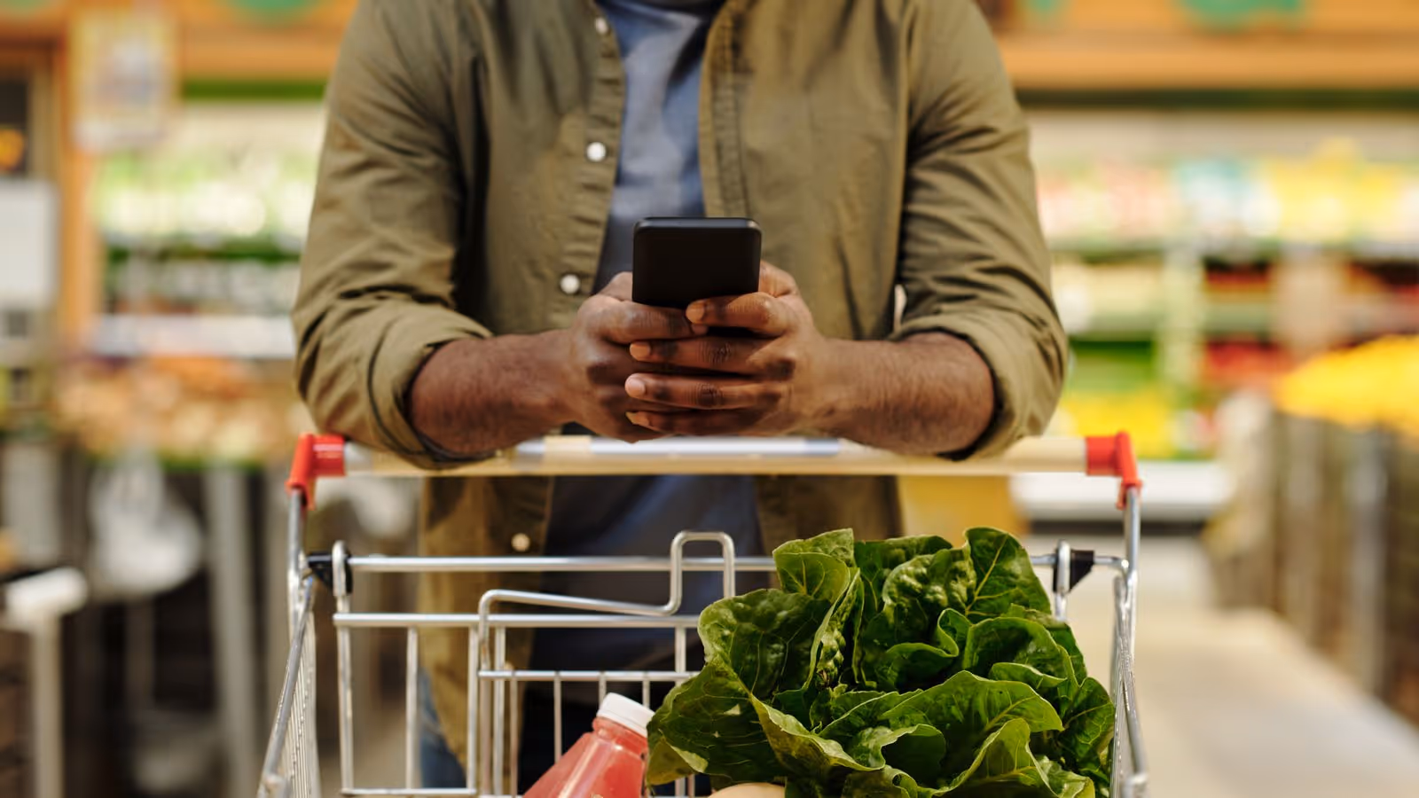 Man grocery shopping while checking his smartphone, with fresh vegetables in a cart, representing healthy food choices and nutrition that support naturally boosting testosterone levels in men.