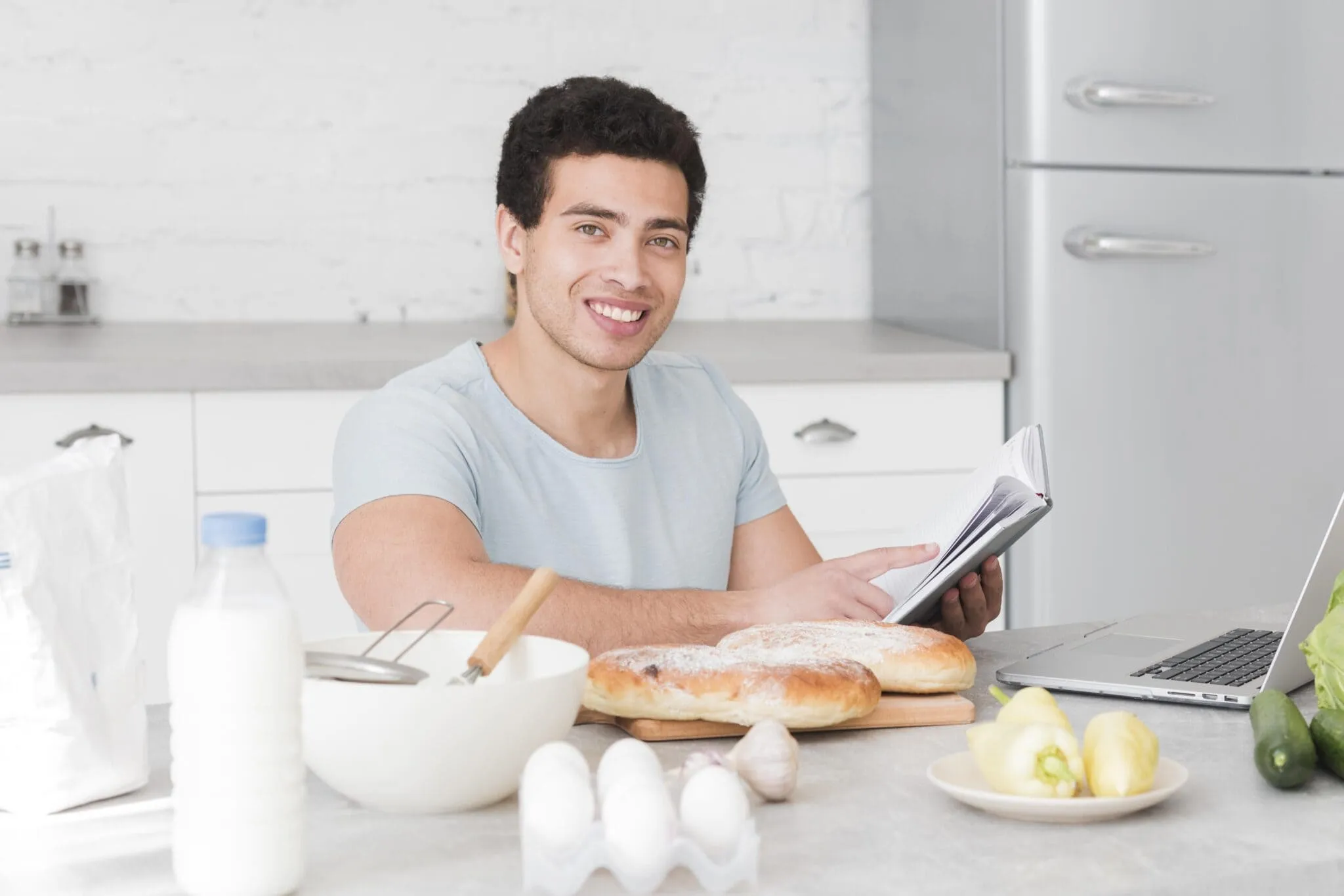 Smiling man reading a recipe book in the kitchen surrounded by bread, eggs, and vegetables, representing awareness of foods that may lower testosterone.