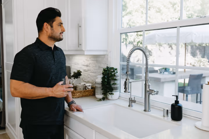 Man standing in a modern kitchen holding a glass of water and looking out the window, representing improved wellness and quality of life associated with shockwave therapy for erectile dysfunction.