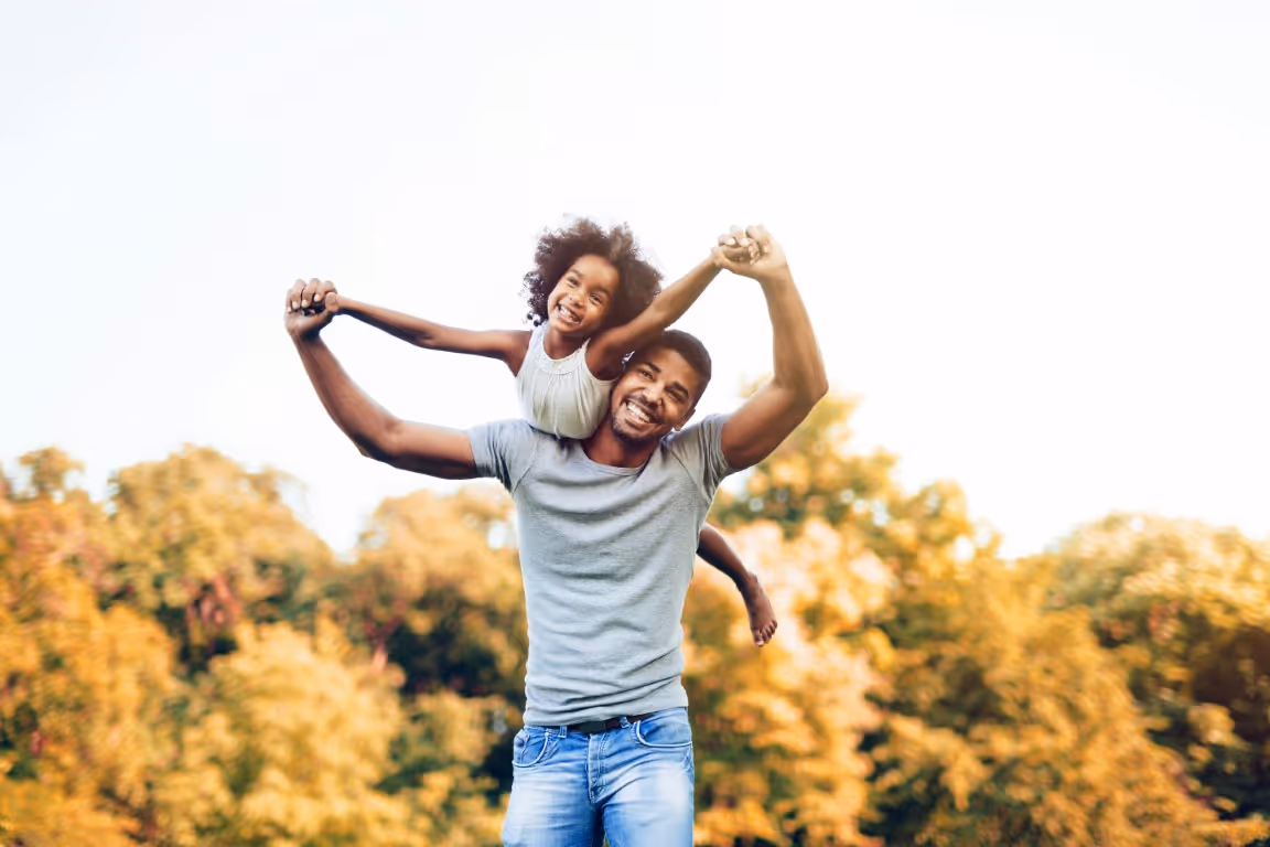 Father smiling while carrying his young daughter on his shoulders outdoors, representing improved energy with testosterone replacement therapy solutions for men.