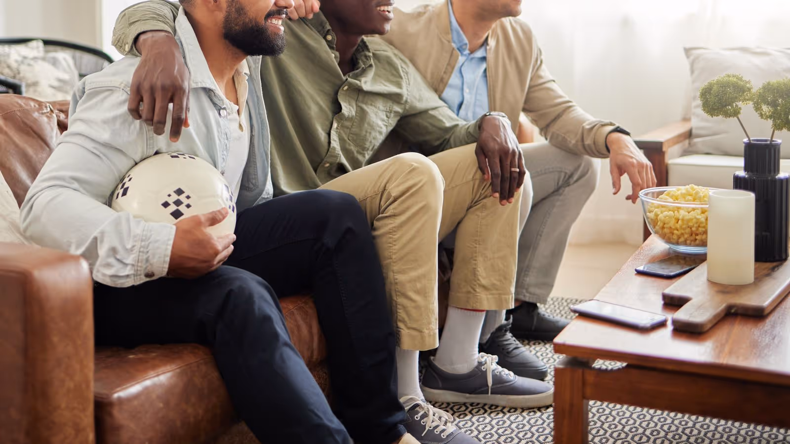 Three friends on a sofa watching TV, one holding a soccer ball, cozy vibe.