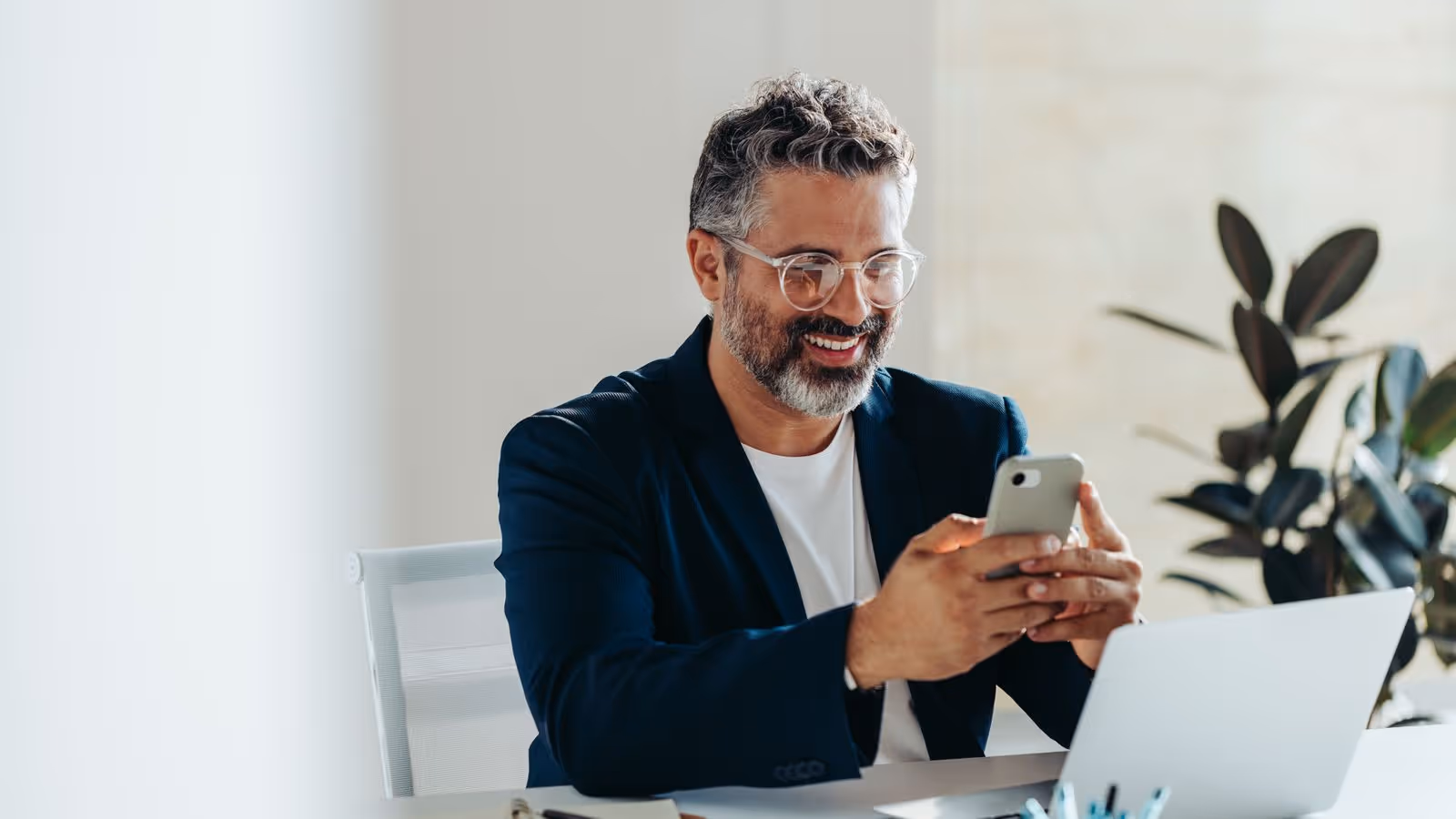 Smiling bearded man with glasses looks at smartphone in bright office.