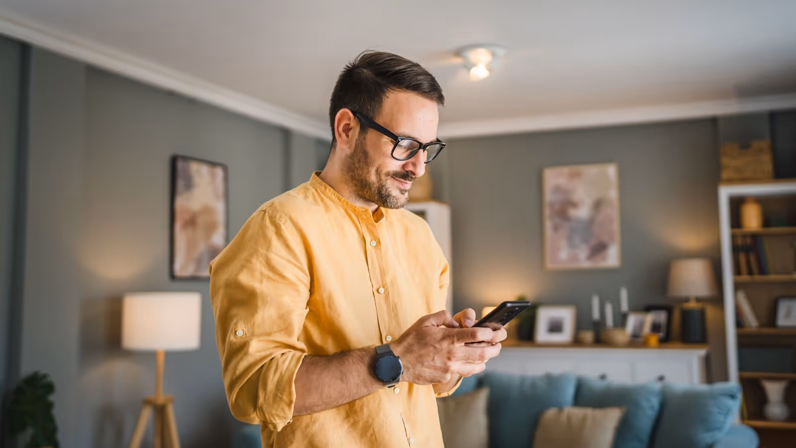 Man in yellow shirt smiling while using smartphone in cozy living room.