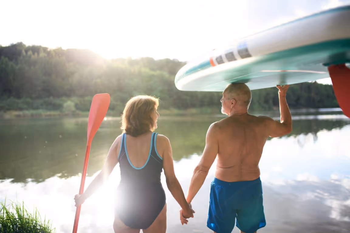 Active older couple holding hands by a lake while carrying paddleboards, symbolizing improved intimacy, vitality, and quality of life supported by effective ED treatment options.