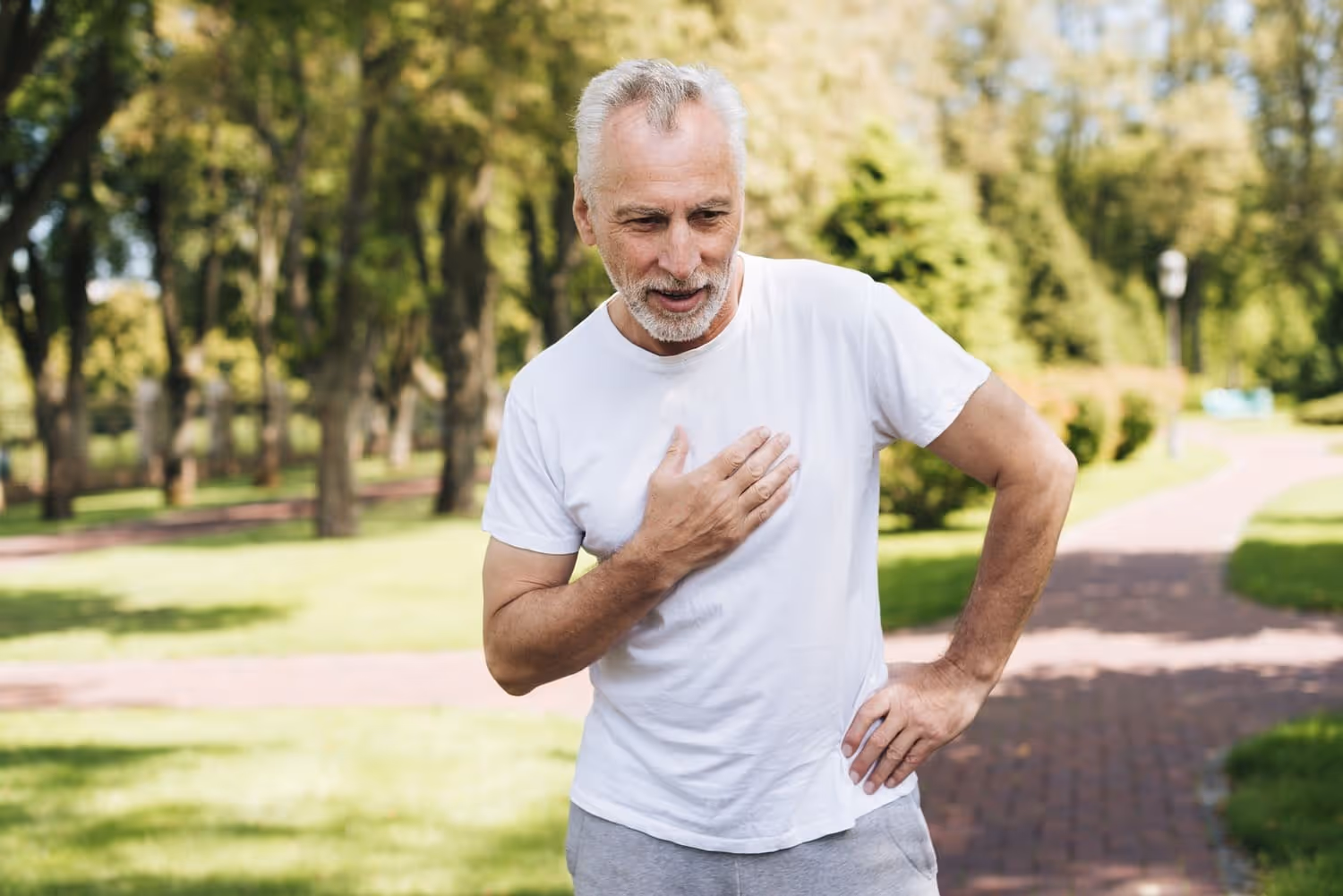 Senior man holds his chest on a park path, reflecting the link between ED and heart health.