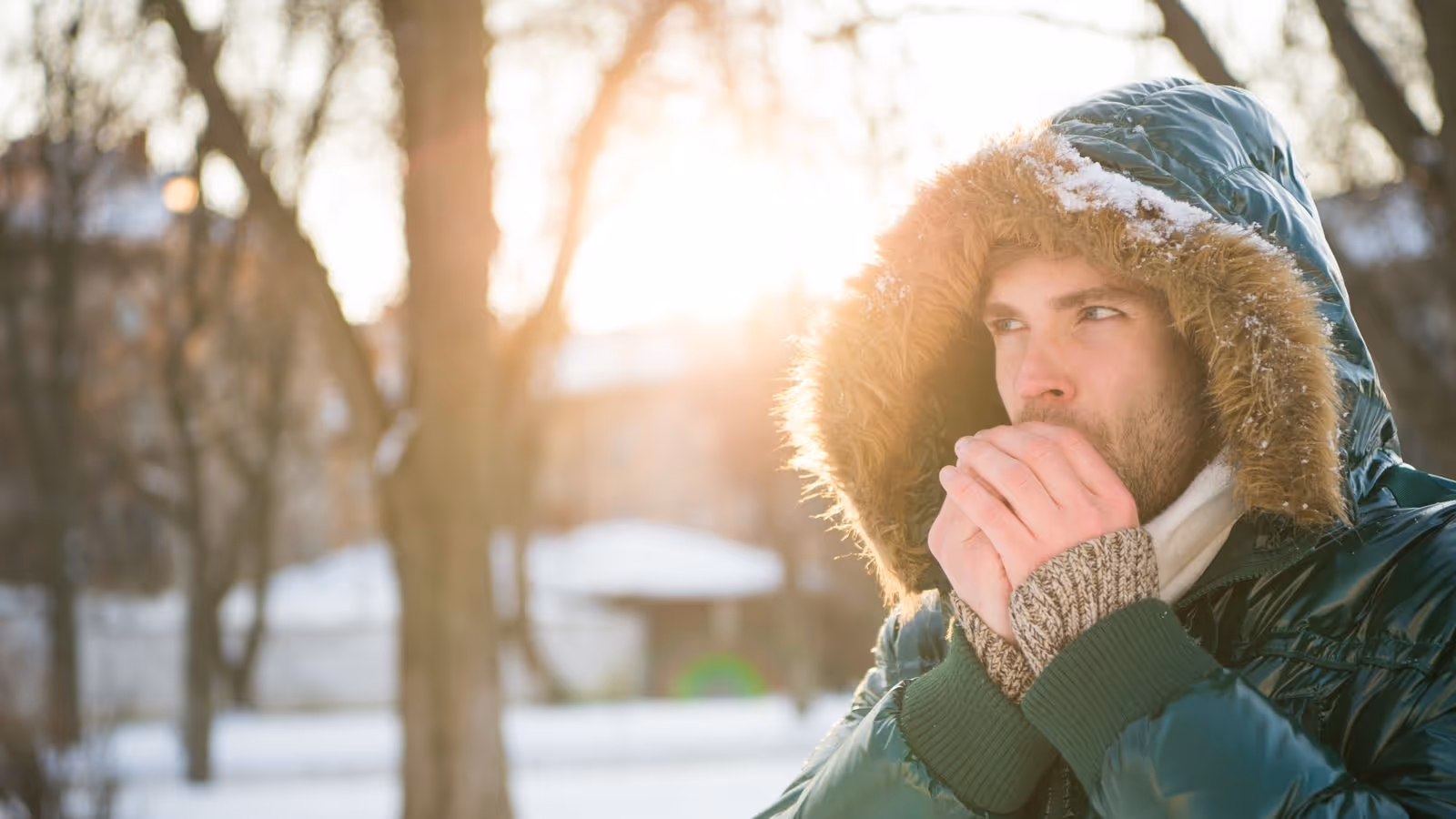 Person in hooded coat warms hands in snowy, sunlit park.