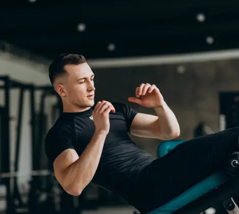 Man in black shirt doing sit-ups on gym bench, dim gym with blurred equipment, showing focus and determination.