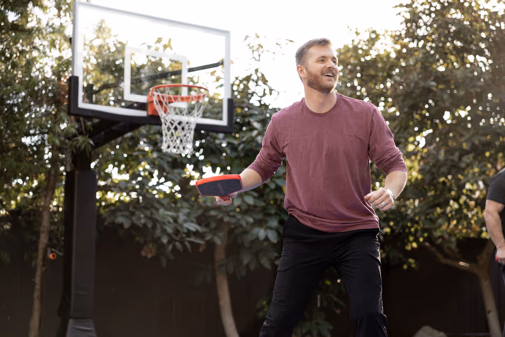 Man in maroon shirt holds paddle near outdoor basketball hoop.