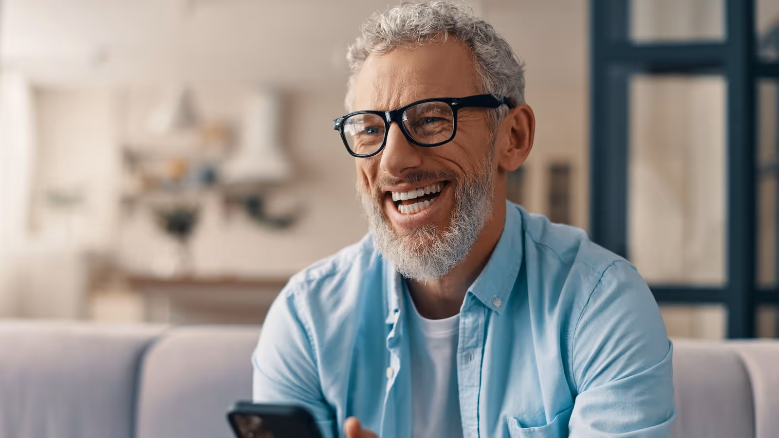 Older man with gray hair and beard laughing while holding a smartphone in a bright room.