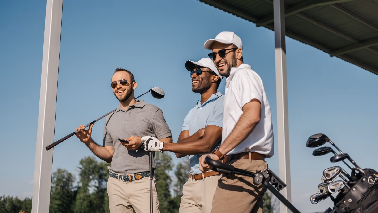 Three men in golf attire smile with clubs at a sunny driving range.