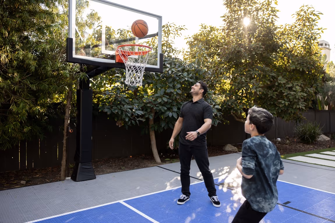 Man and boy play basketball outside on a sunny court surrounded by trees.