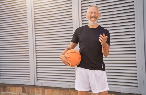 Elderly bearded man smiling, holding a basketball, wearing black shirt and white shorts, standing by a metal wall.
