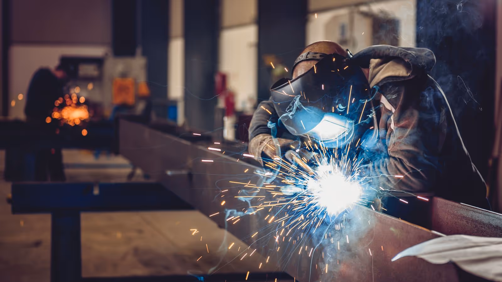 Welder in protective gear works with sparks flying in workshop.