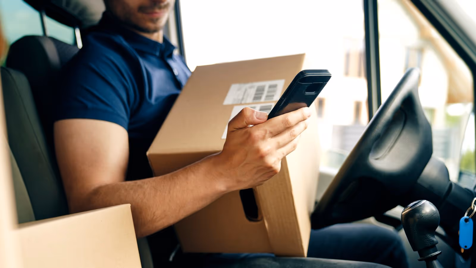 Delivery worker holding a package and using a phone in a van.