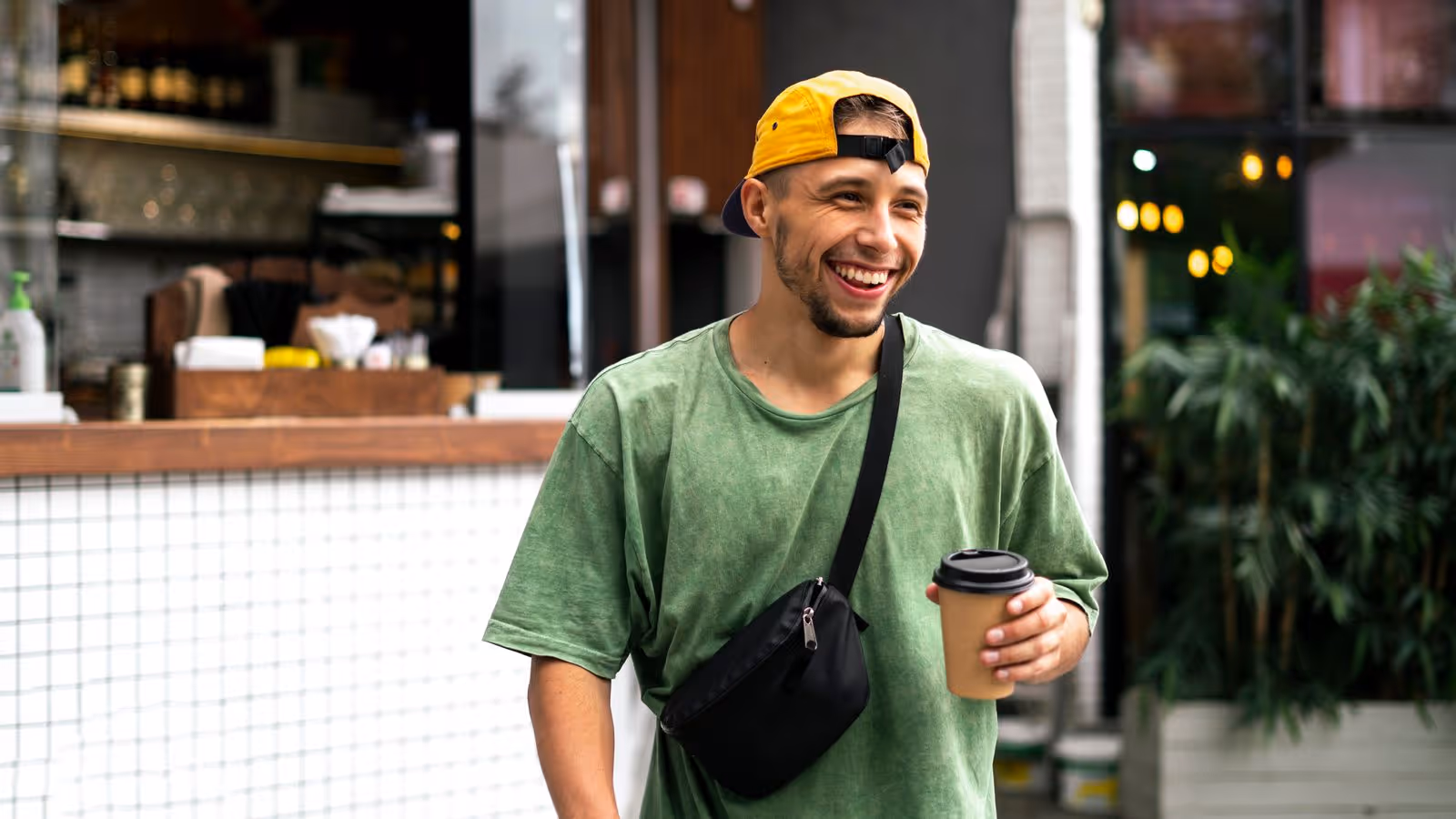 Smiling man in yellow cap holds coffee outside café, reflecting vitality—clomid therapy.