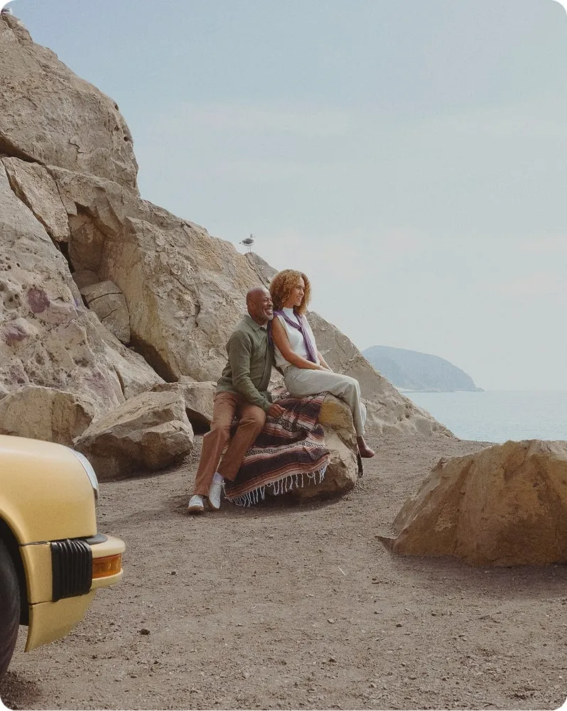 Happy couple sitting on a large rock by the ocean, smiling and enjoying each other’s company, symbolizing the restored intimacy and sexual confidence possible with PT-141 therapy.