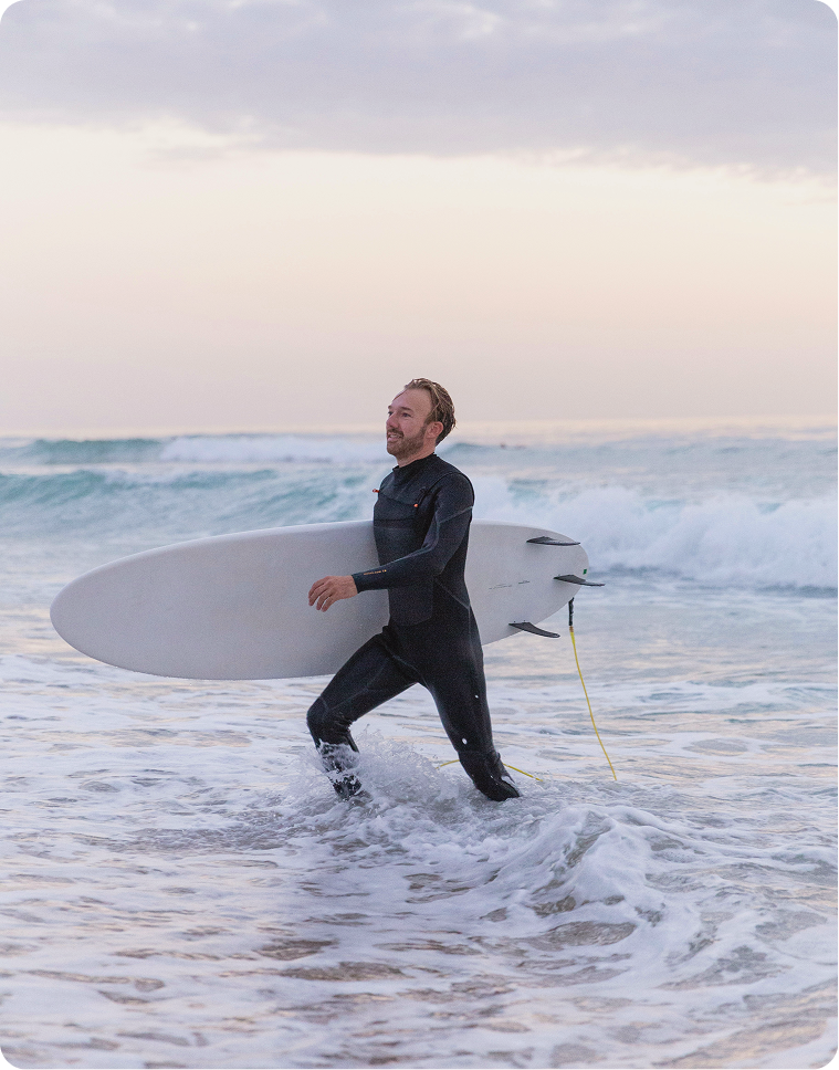 Man in a wetsuit carrying a surfboard at the ocean, representing Gameday Men’s Health testosterone cypionate injection therapy focused on energy, strength, and performance