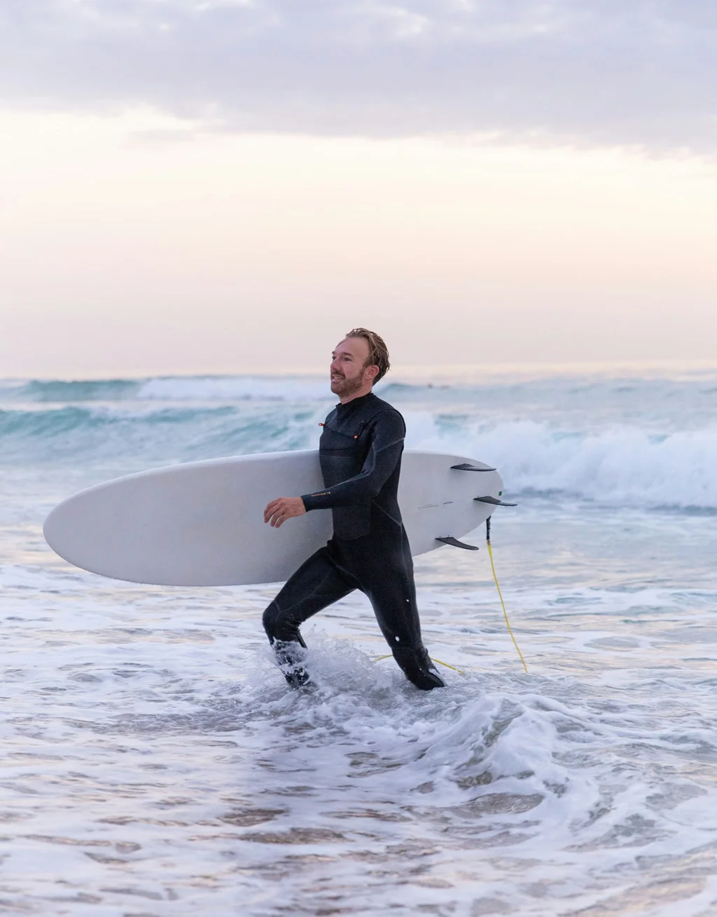 Man in a wetsuit carrying a surfboard at the ocean, representing Gameday Men’s Health testosterone cypionate injection therapy focused on energy, strength, and performance