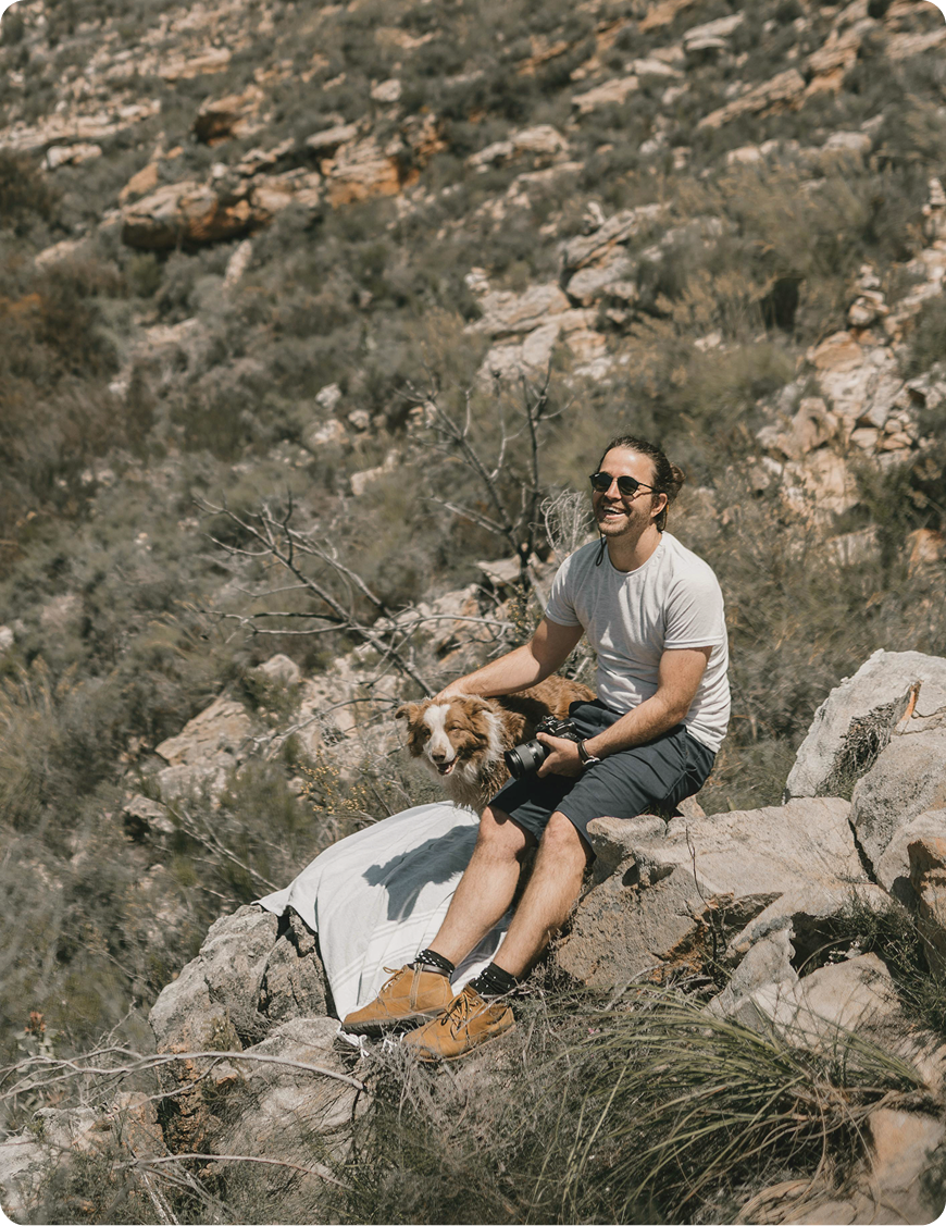 Smiling man resting in a mountain setting with his dog and camera, capturing a moment of adventure and personal fulfillment aligned with Vitamin B Complex Injections in West Edmonton, AB.