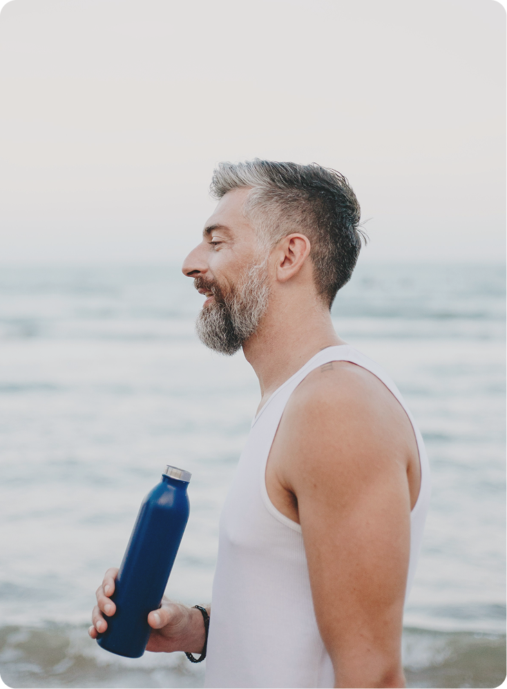 Happy man walking along the beach while drinking water, reflecting confidence and daily routine supported by Minoxidil + Finasteride in Ontario at Gameday Men’s Health