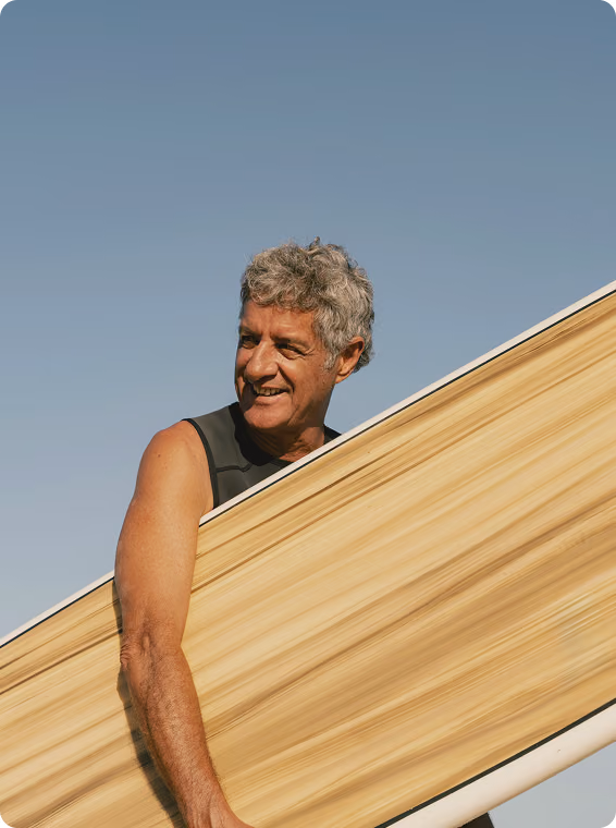 Older man carrying a surfboard toward the beach, reflecting active aging and resilience associated with Vitamin C Injections in El Paso