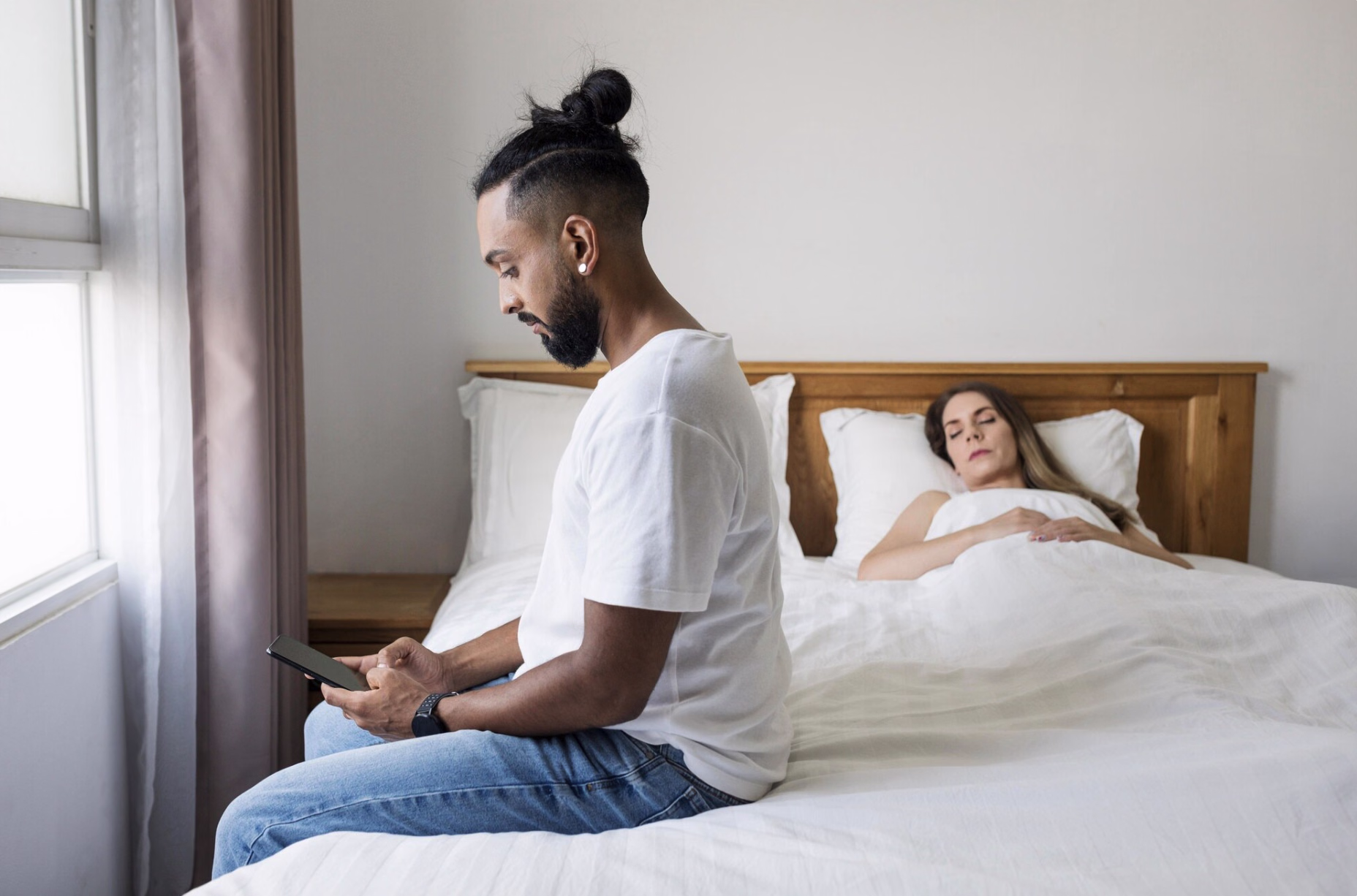 Sad, thoughtful man sitting on the edge of the bed looking at his phone while his partner lies nearby, illustrating the emotional impact of erectile dysfunction and hormone imbalance.