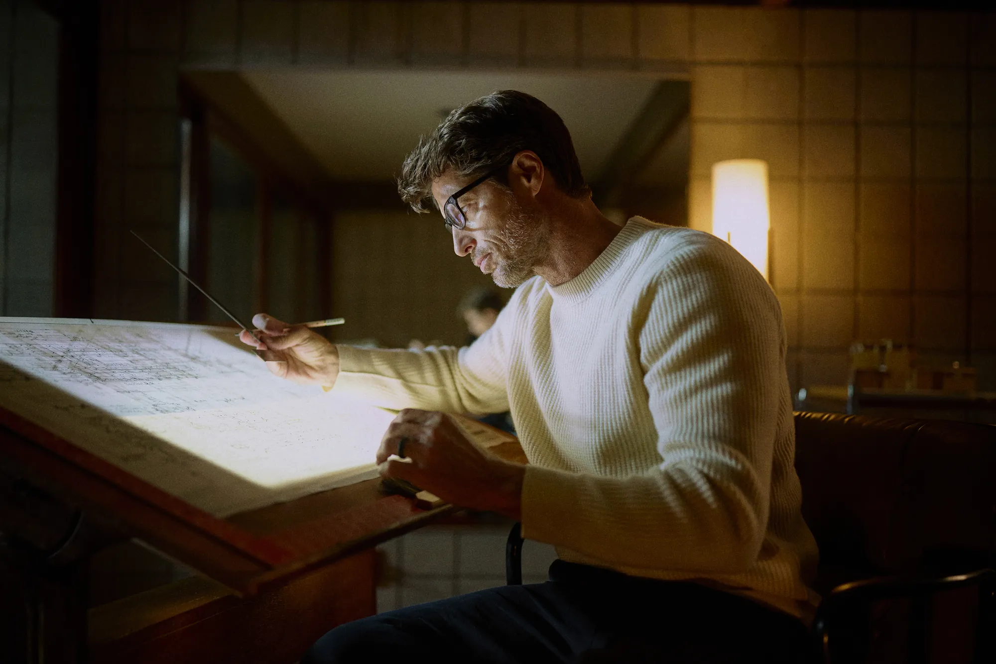 Man in glasses working late at a drafting table under lamp light