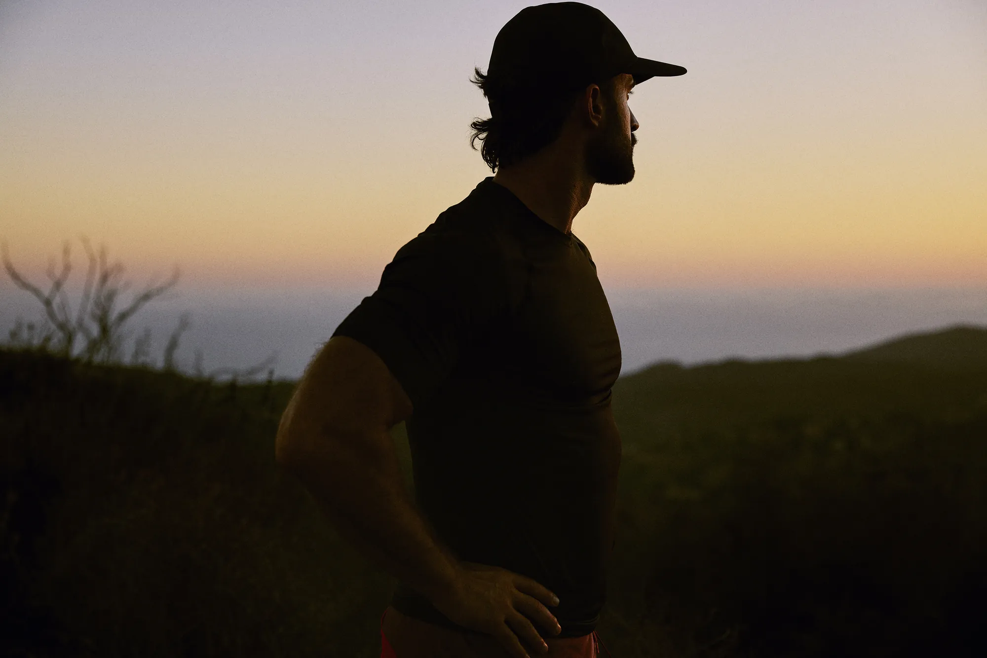 Man in a ball cap standing on a hillside trail at sunset