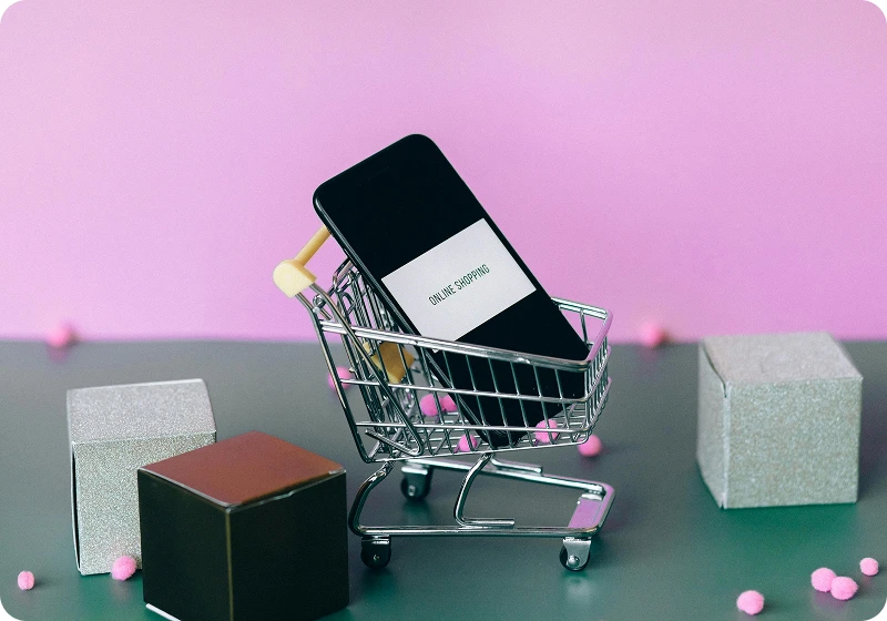 Smartphone with label 'ONLINE SHOPPING' placed inside a miniature shopping cart on a table with decorative cubes and pink pom-poms against a pink background.