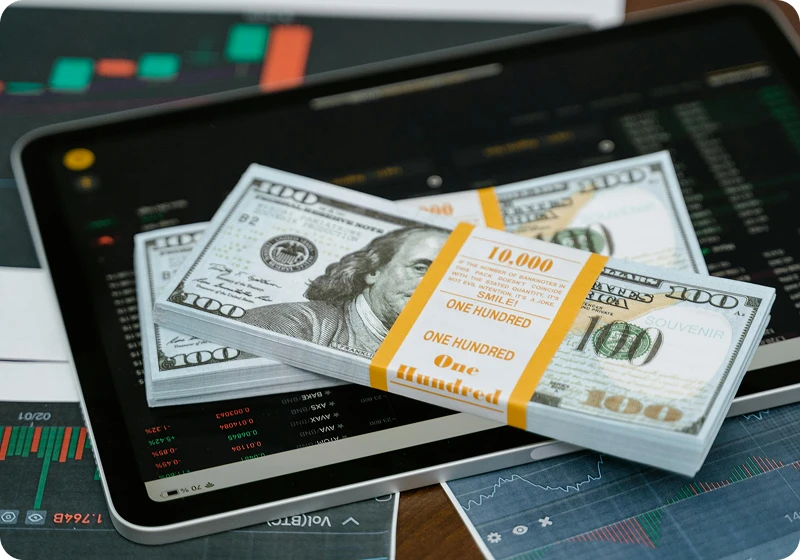 Stacks of US one hundred dollar bills placed on two tablet devices showing financial charts and stock market data.