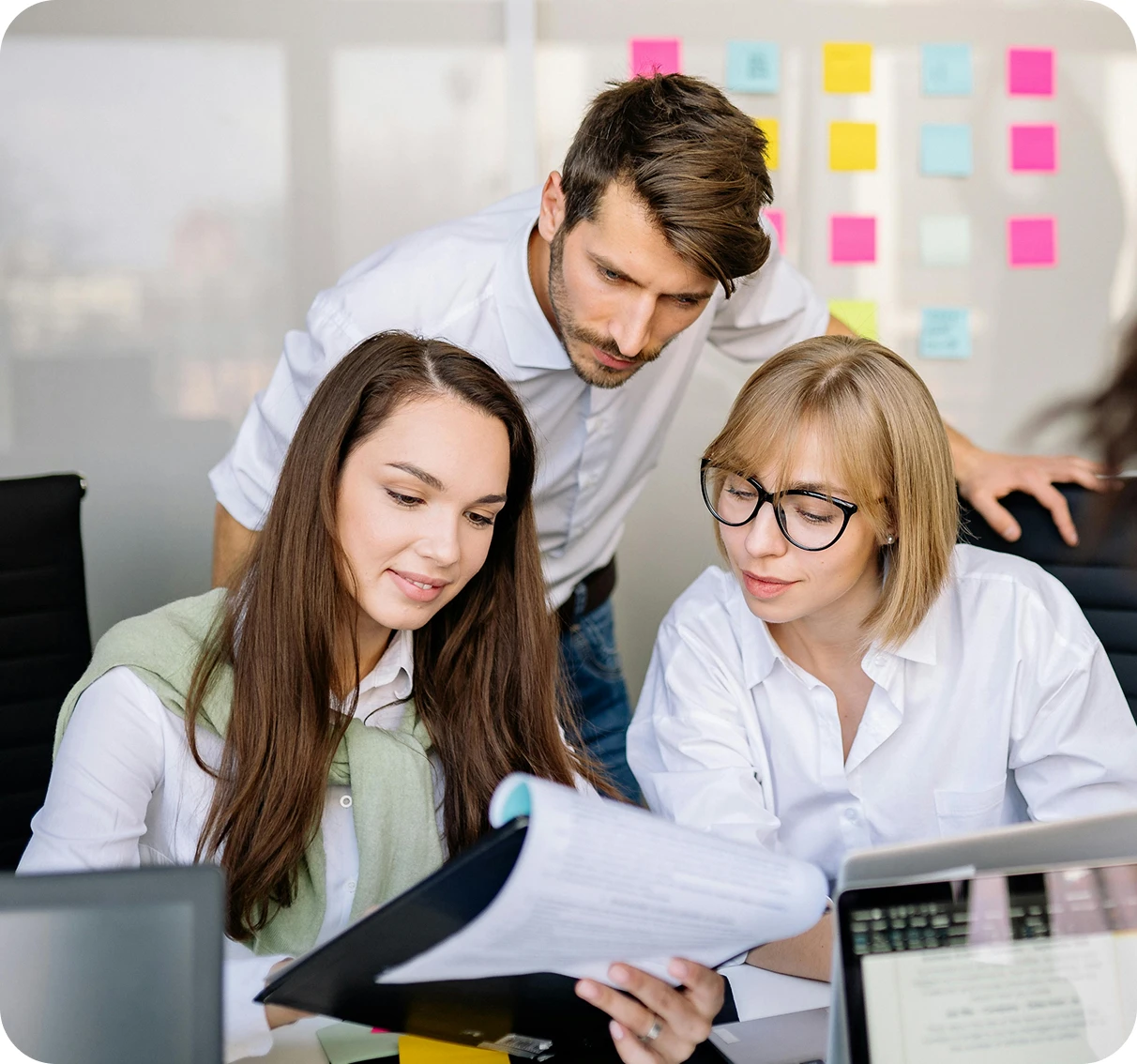 Three colleagues in a modern office reviewing documents together, with colorful sticky notes on the wall in the background.