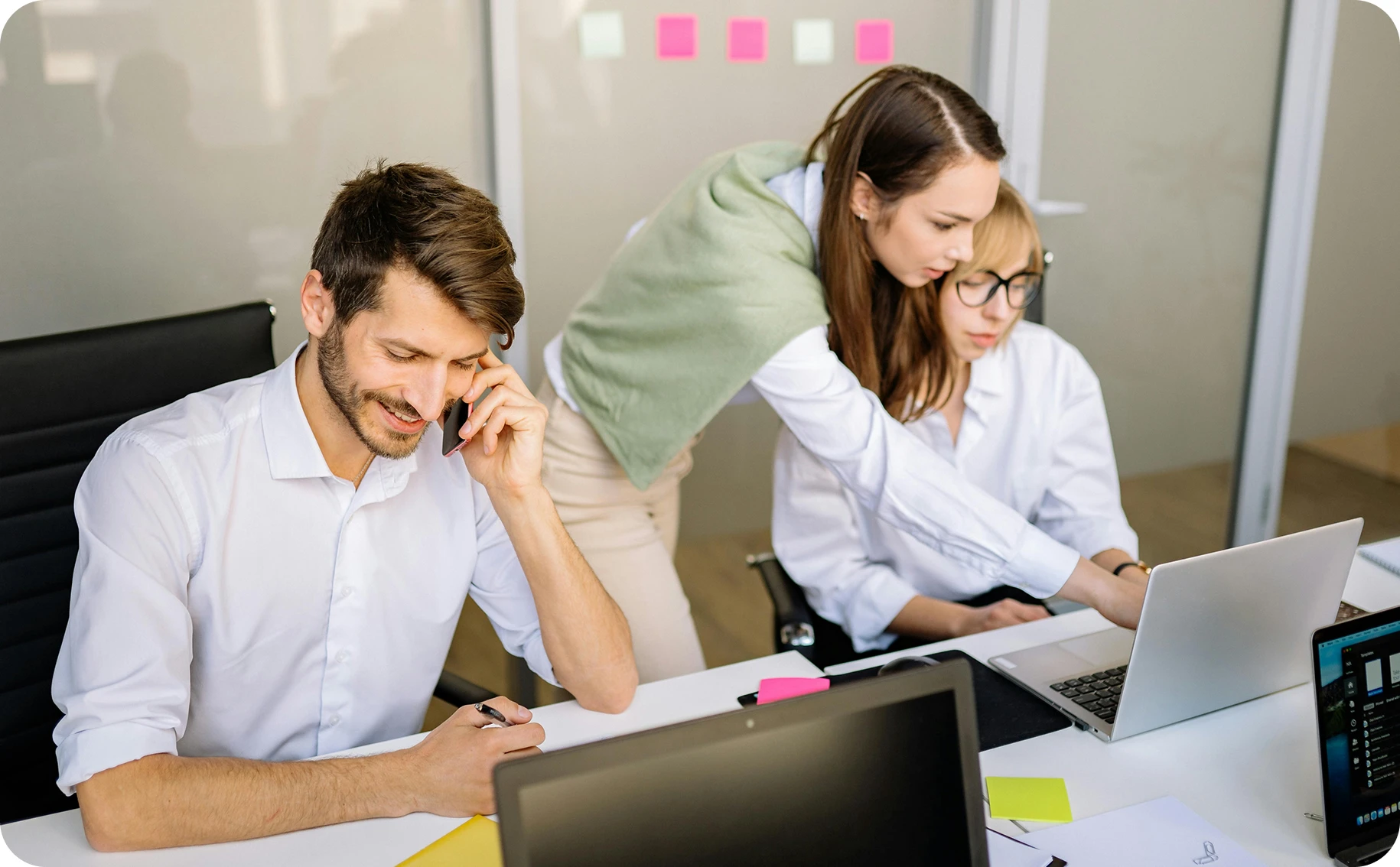 Three young professionals working together in an office, with one man on the phone and two women collaborating over a laptop.