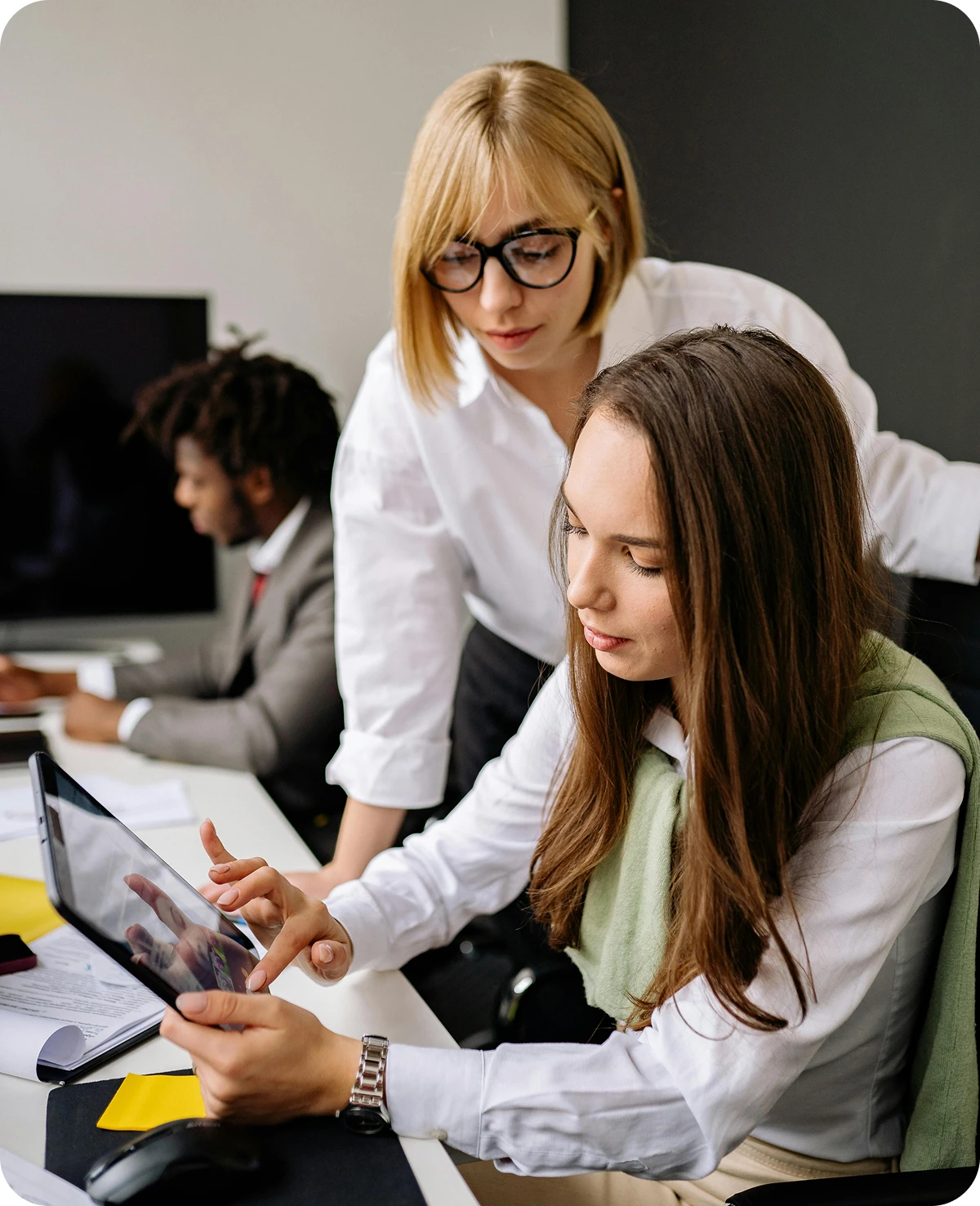 Two women working together using a tablet in an office, with a man in a suit working at a computer in the background.