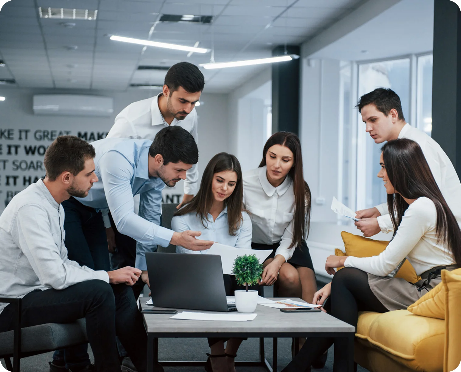 Group of six young professionals collaborating around a table with laptop and documents in a modern office.